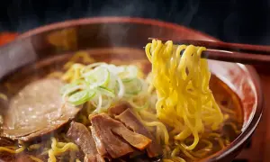 En skål med ångande ramen, med en skillnad på nudlar och pasta som tydligt syns. Nudlarna lyfts med ätpinnar, med kött och grönsaker i buljongen.