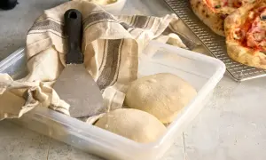 Pizzadeg redo att baka ut. En degskrapa och en kökshandduk ligger bredvid en plastlåda med två degbollar.