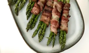 Grön sparris insvept i knaprigt bacon, serverad på en ljusblå tallrik. En utsökt kombination av grönsaker och baconlindad sparris.