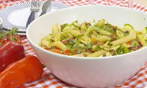 En stor skål med vegansk pastasallad med penne, zucchini, tomater och oliver. Serveras på en rödvitrutig duk.