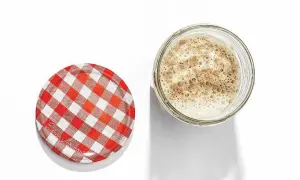 En bubblig surdeg i ett glasburk, bredvid ett rött och vitt rutigt lock. Så här ska en surdeg se ut när den är aktiv och redo att bakas med.
