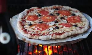 En pizza med tomater, champinjoner och oliver grillas över glödande kol. Perfekt för den som vill testa att frysa pizzadeg och baka utomhus.