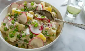 En krämig grekisk potatissallad med rädisor och gräslök, serverad i en vit skål med en sked.