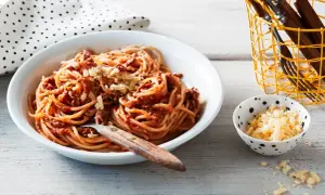 En skål med stuvad spaghetti med köttfärssås och riven ost. En liten skål med mer ost och en korg med bestick står bredvid.