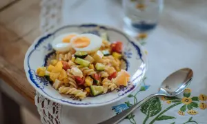 En färgglad pastasallad med ägg, grönsaker och ost, perfekt som pastasallad barn. En sked och ett glas vatten står bredvid.
