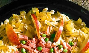 En närbild av en pastasallad med lufttorkad skinka, äppelklyftor och gröna ärtor.