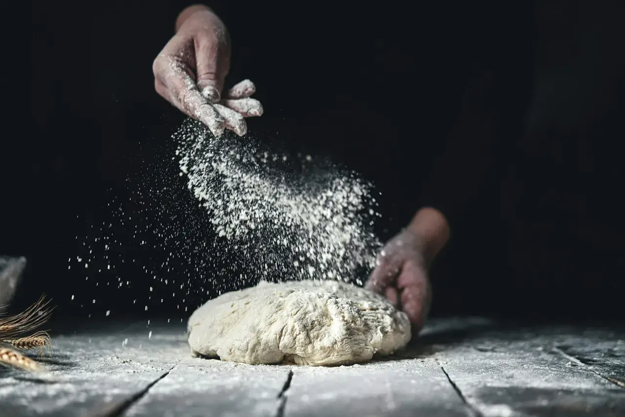 Händer strör mjöl över en deg. Det ser ut som en svensk hemmabakare som förbereder sitt bröd med fint **bread flour svenska**.