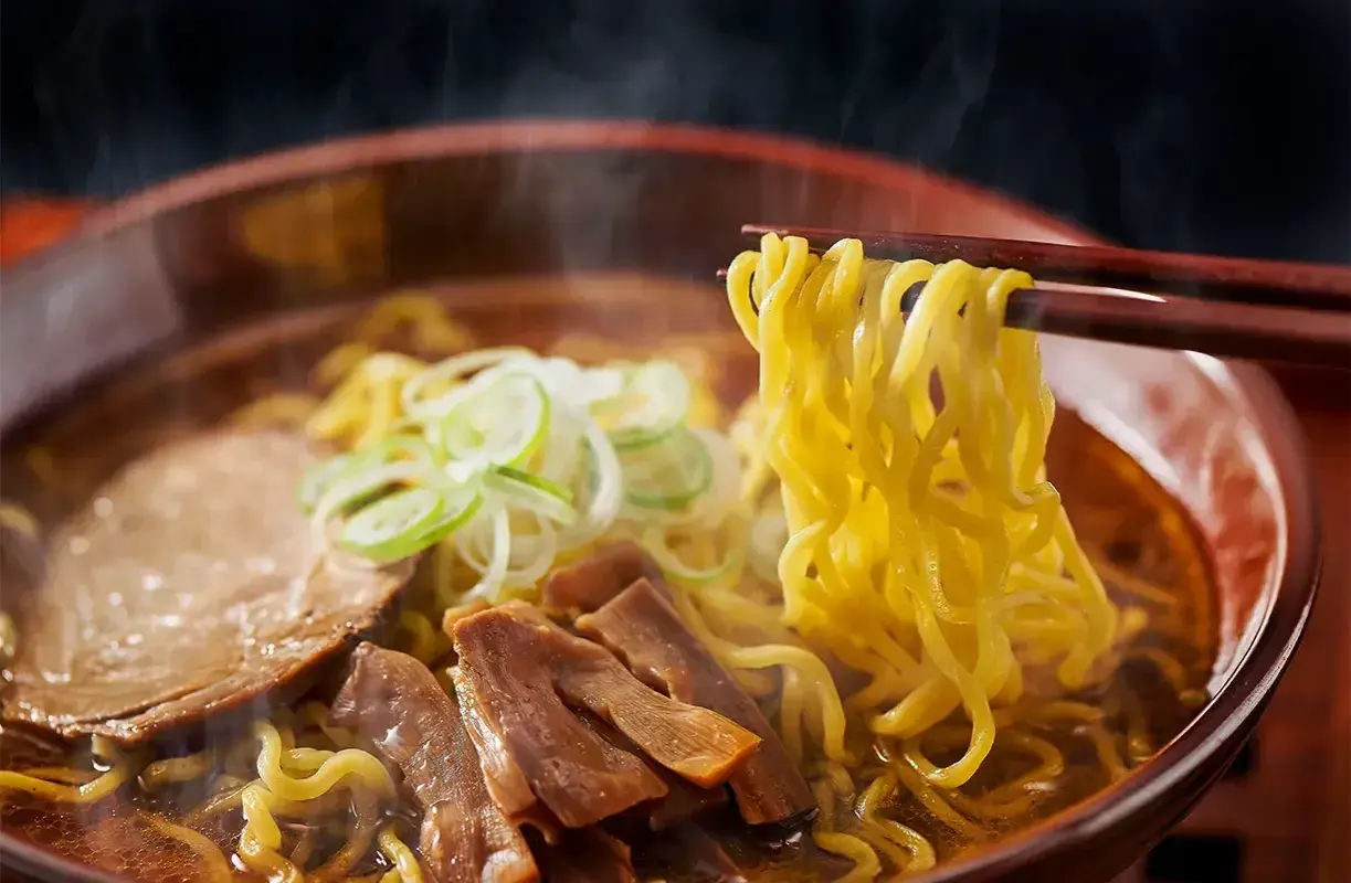 En skål med ångande ramen, med en skillnad på nudlar och pasta som tydligt syns. Nudlarna lyfts med ätpinnar, med kött och grönsaker i buljongen.