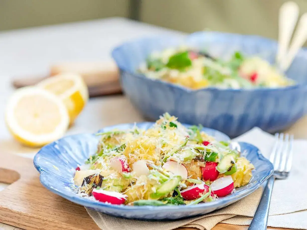 En härlig potatissallad med sparris, rädisor och hyvlad ost. Serveras på en blå tallrik med citroner i bakgrunden.