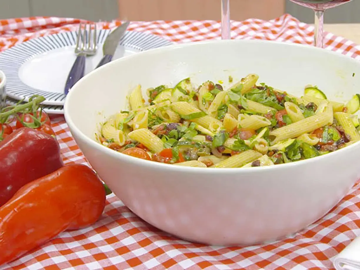 En stor skål med vegansk pastasallad med penne, zucchini, tomater och oliver. Serveras på en rödvitrutig duk.