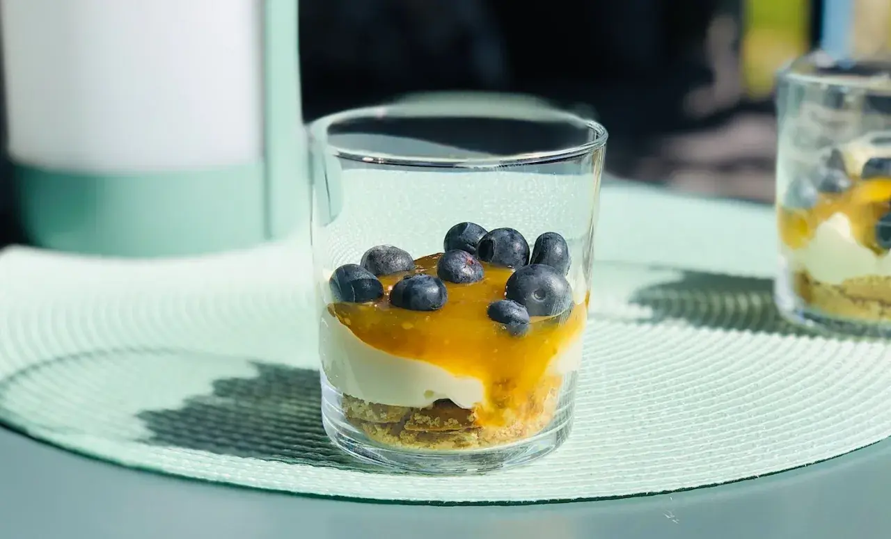 En läcker cheesecake i glas med blåbär och en gyllene sås. Perfekt för en solig sommardag.