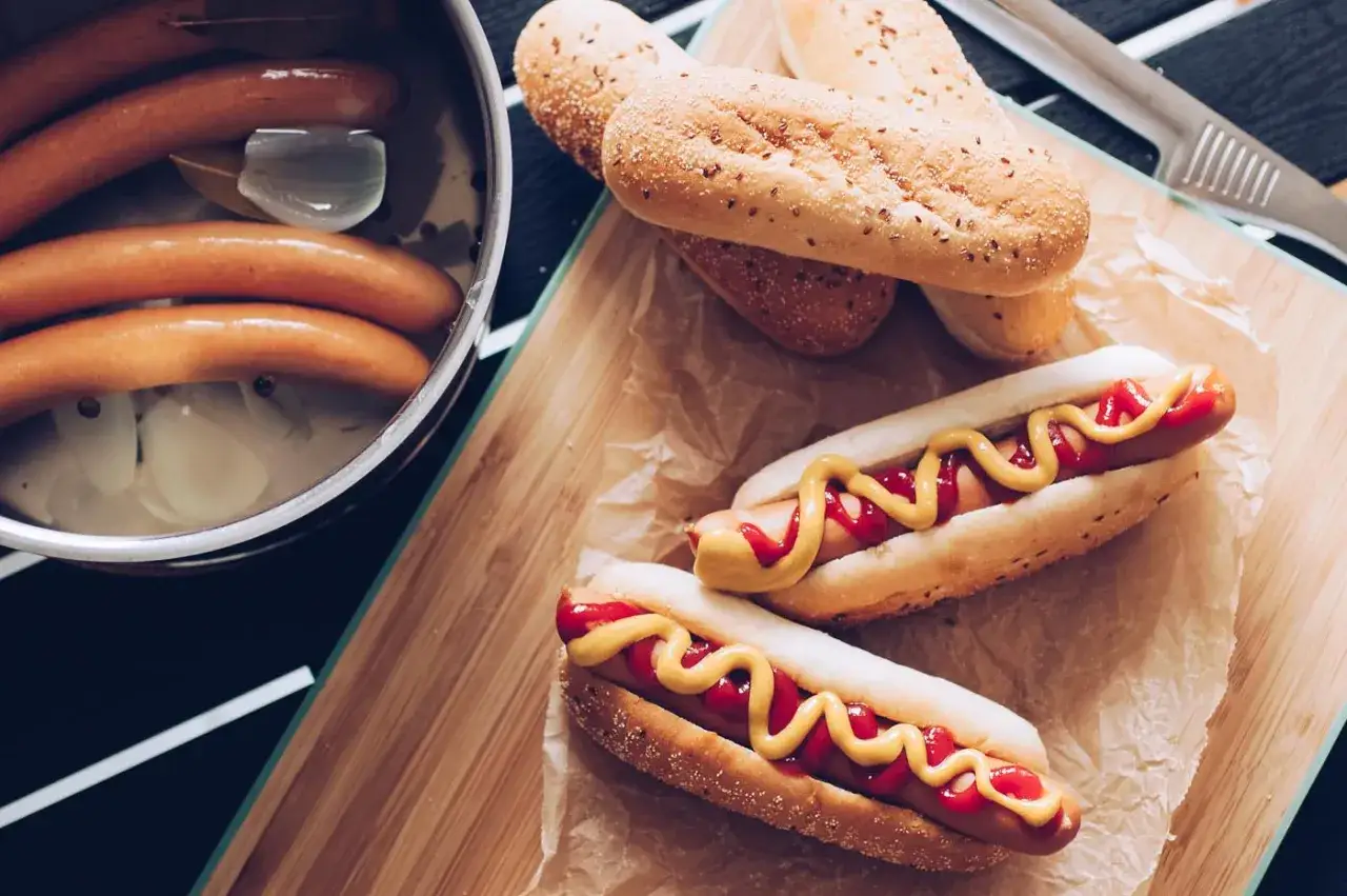 Här ser du hur kokar man korv. Korvarna ligger i en kastrull med vatten och lök. Bredvid ligger korvbröd och färdiga korvar med senap och ketchup.