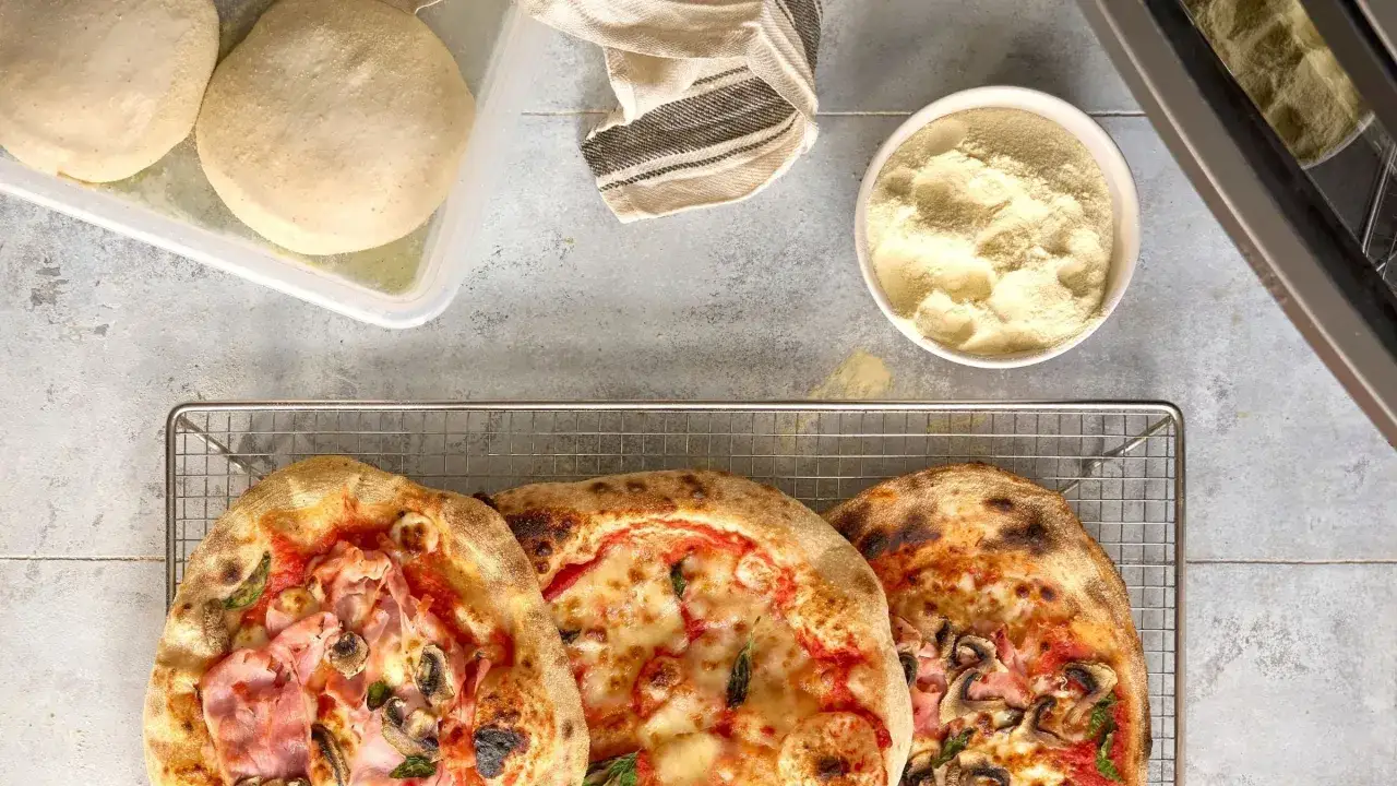 Tre färdiga pizzor på ett galler, redo att avnjutas. Bredvid ligger pizzadeg och en skål med mjöl, redo att baka ut pizza.