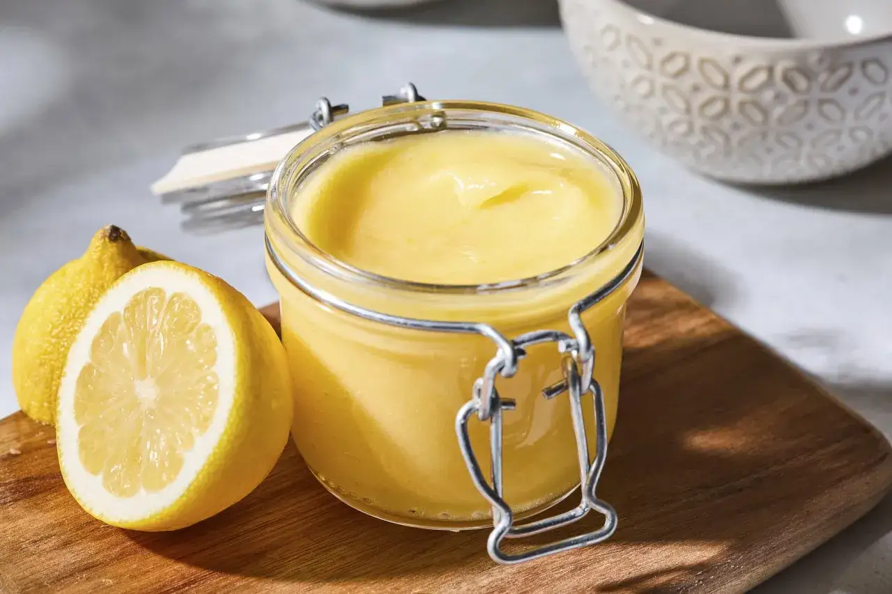 En burk med krämig lemon curd står bredvid färska citroner på en träskärbräda.