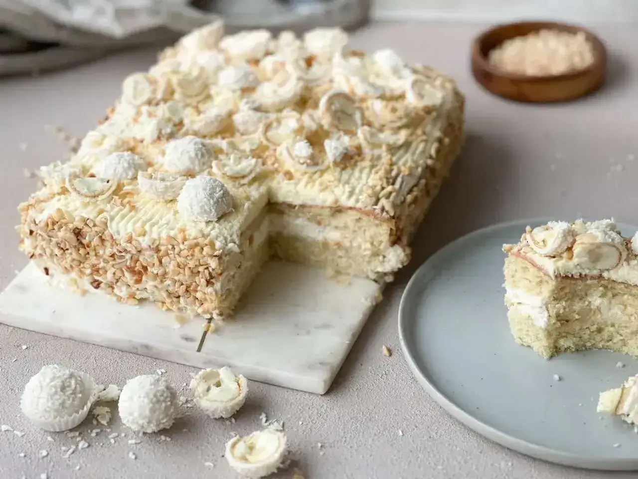 En vit, krämig tårtbotten långpanna dekorerad med nötter och chokladpraliner. En bit ligger på en tallrik.