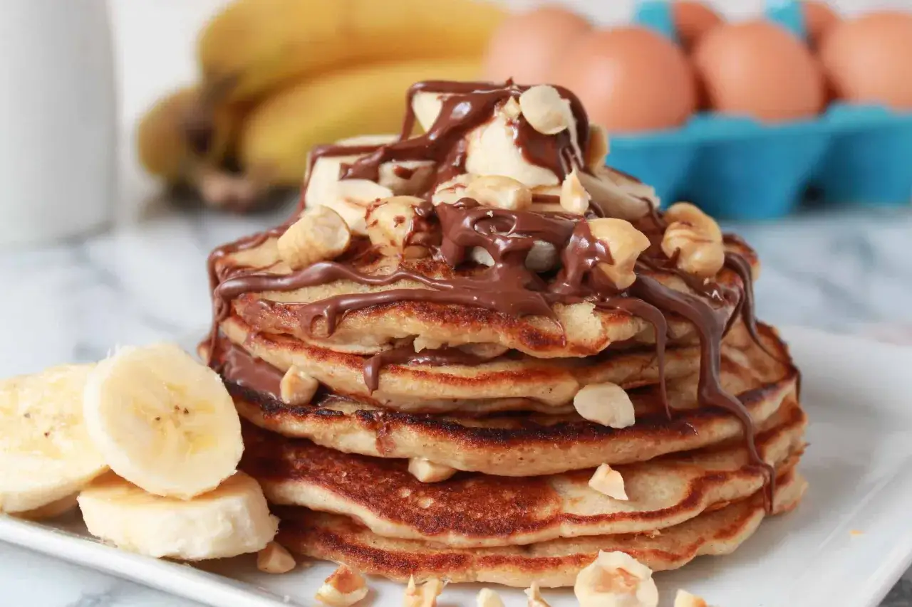 En hög med pannkakor toppad med banan, hasselnötter och rikligt med nutella. En dröm för alla som älskar pannkakstårta med nutella!