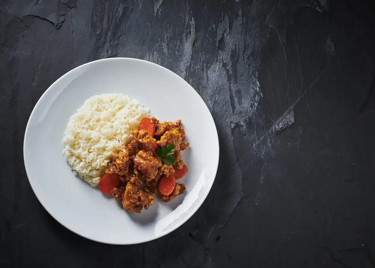 En tallrik med ris och krispig orange chicken, garnerad med persilja och morotsbitar. En läcker måltid som ser ut att vara redo att avnjutas.