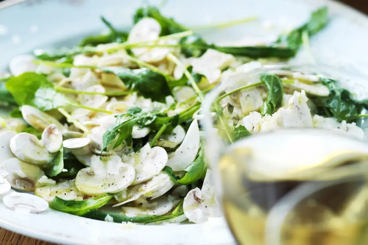 En härlig sallad med marinerade champinjoner, färsk spenat och ruccola, serverad på en ljusblå tallrik. Ett glas vitt vin skymtar i förgrunden.