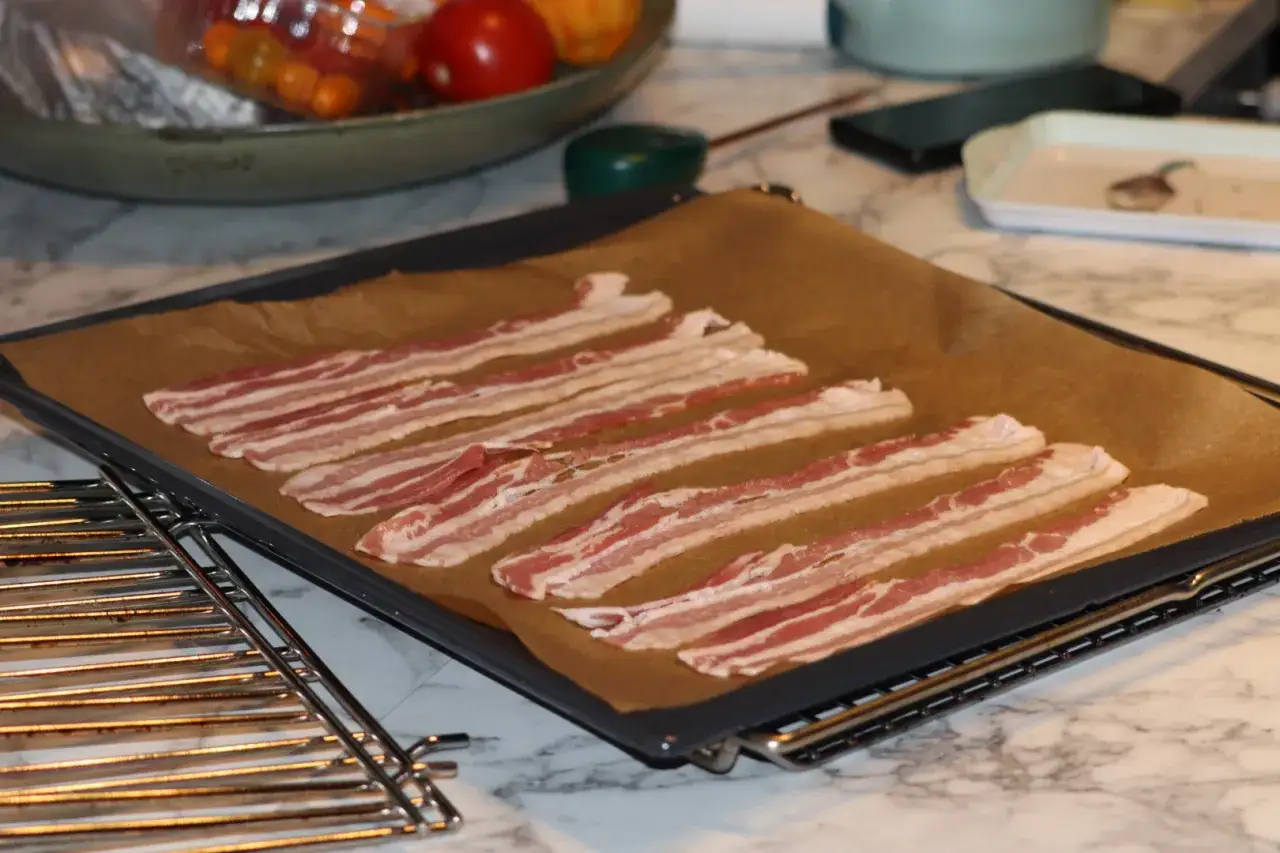 Råa baconskivor ligger utspridda på ett bakplåtspapper, redo för att tillagas i ugnen. En härlig start på en god måltid.