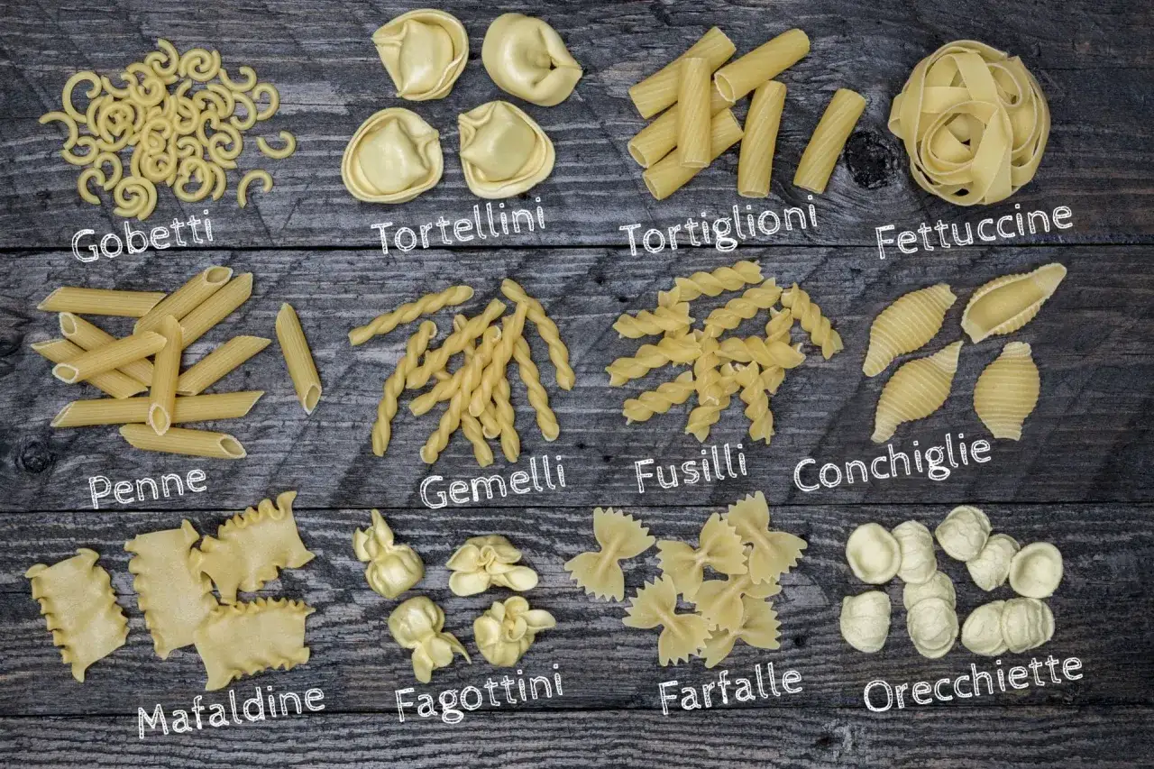 En mängd olika pasta typer, som penne, fusilli och farfalle, ligger utspridda på en träbakgrund.