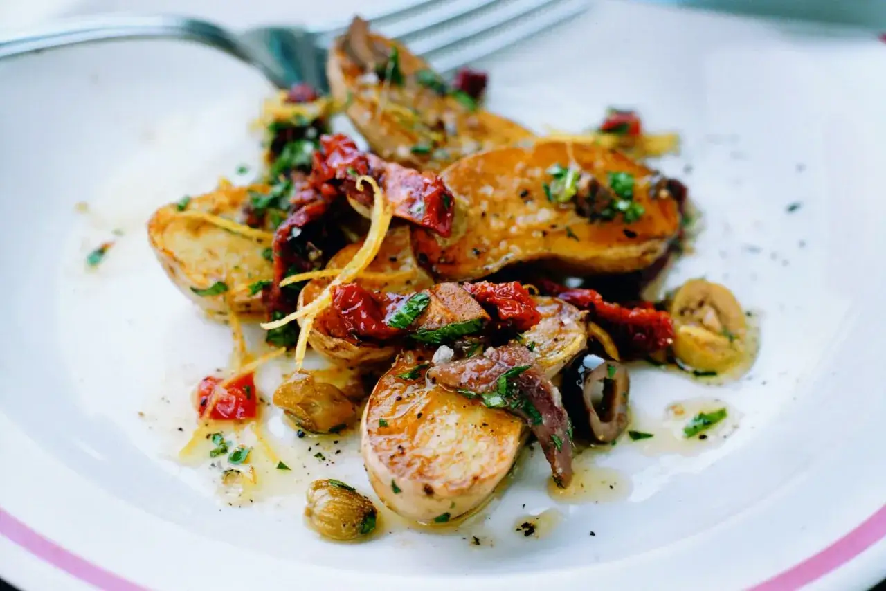 En tallrik med gyllene, stekt potatis toppad med soltorkade tomater, oliver och färska örter. En läcker rätt som ser ut att vara perfekt tillagad.