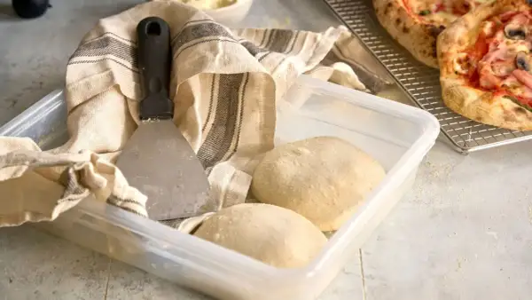 Pizzadeg redo att baka ut. En degskrapa och en kökshandduk ligger bredvid en plastlåda med två degbollar.